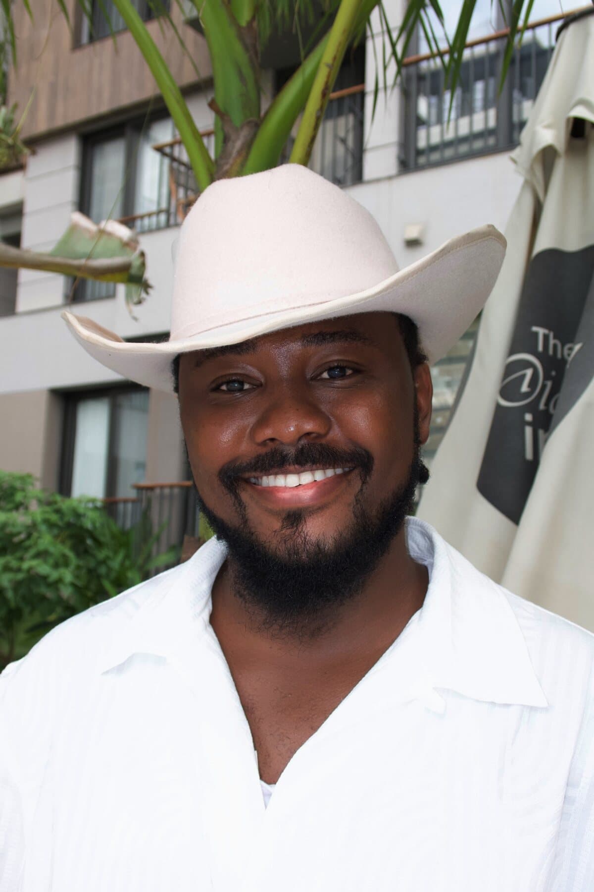 Warm smiling portrait of ABJKOREA (Victor) in a cream hat and white shirt against tropical greenery.