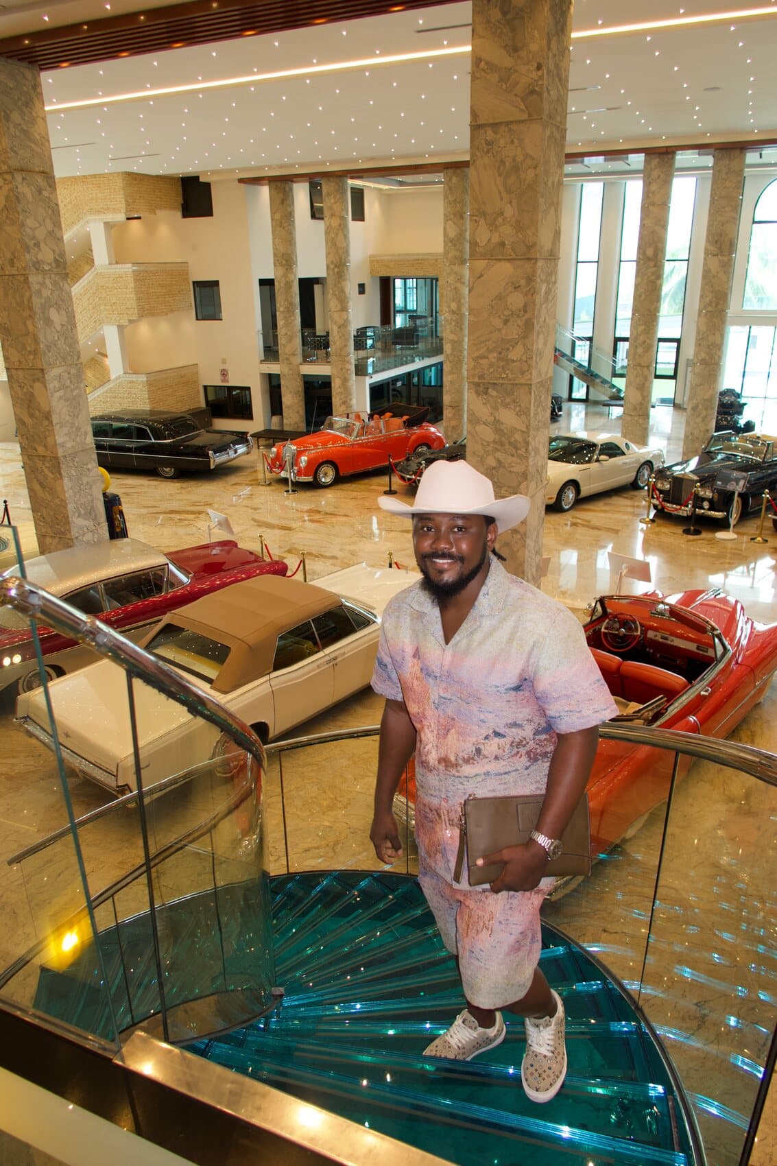 ABJKOREA descending a glass staircase above a collection of classic automobiles.